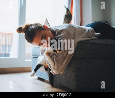 Side view of young lady in casual clothes lying down on comfortable couch playing with adorable calico cat in modern apartment Stock Photo