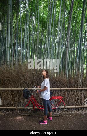 Attractive young caucasian Woman with her bicycle looking up at trees in landmark Arashiyama Bamboo Grove forest in Kyoto, Japan Stock Photo