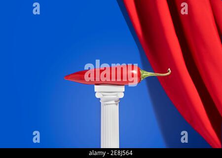 Red hot pepper placed on miniature column against blue background with red velvet curtain Stock Photo
