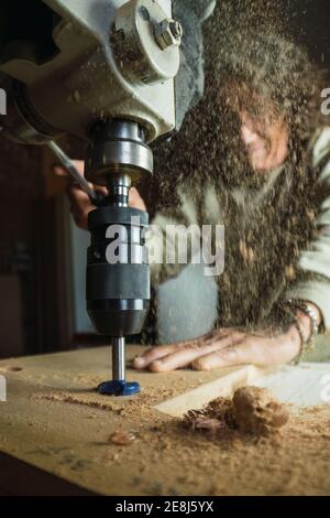 Low angle of male carpenter without a thumb drilling hole in wooden plank while working in dusty carpentry workshop Stock Photo