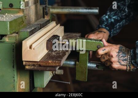 Crop unrecognizable tattooed male carpenter drilling wooden board using lathe during work in joinery Stock Photo
