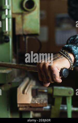 Crop unrecognizable tattooed male carpenter drilling wooden board using lathe during work in joinery Stock Photo