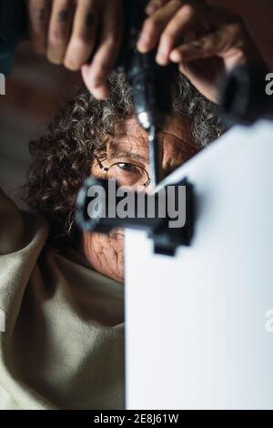 Concentrated unrecognizable senior male furniture maker with curly hair using screwdriver while fixing plinth legs to cabinet in workshop Stock Photo