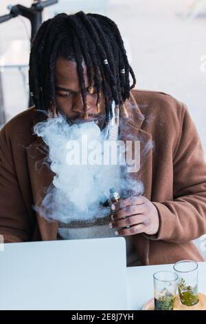 Focused adult hipster African American businessman with dreadlocks smoking cigarette while working with laptop at table with drinks in outdoor restaur Stock Photo