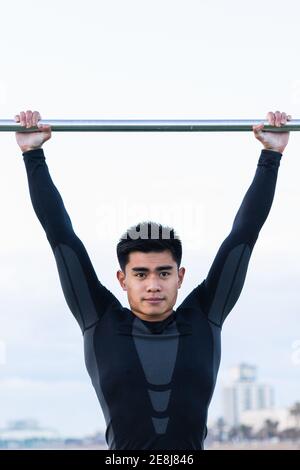 Strong determined young Asian man in sportswear doing exercises on monkey bars during outdoor workout on sports ground Stock Photo