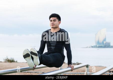 Full body of young Asian male athlete in sportswear doing L Sit exercise on parallel bars during calisthenics workout on sports ground Stock Photo