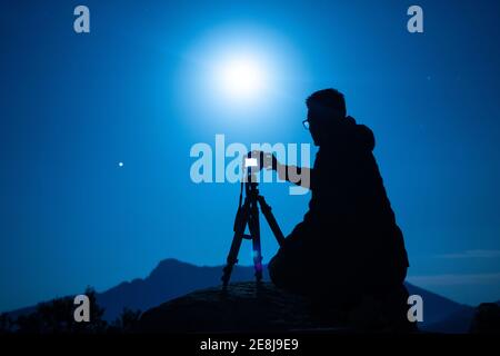 Side view of unrecognizable male traveler silhouette with photo camera on tripod against ridge under blue sky with shiny sun at night Stock Photo