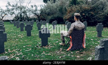 Back view full body of soldier in uniform sitting on chair with American flag while mourning death of warriors at graveyard Stock Photo
