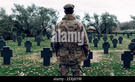 Back view anonymous soldier wearing camouflage outfit standing on vast military cemetery on early autumn day Stock Photo