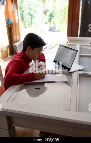 High angle side view of smart schoolboy sitting at table with laptop and writing in notebook while doing homework alone Stock Photo