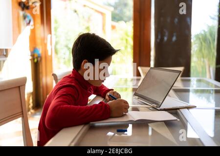 Side view of smart schoolboy sitting at table with laptop and writing in notebook while doing homework alone Stock Photo