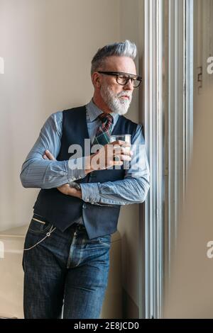 Confident middle aged stylish male with gray hair and beard in trendy outfit and eyeglasses leaning on wall and looking out window while drinking whis Stock Photo