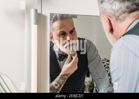 Crop serious middle aged male executive with tattoo touching gray beard while looking in mirror in house Stock Photo