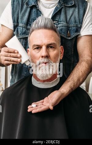 Crop anonymous male barber standing behind serious trendy bearded middle aged client and preparing for fixing haircut with cream Stock Photo