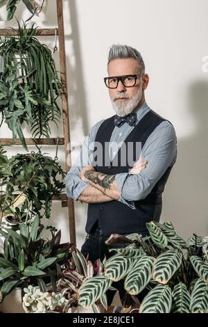 Self assured mature male entrepreneur with tattoo and folded arms looking at camera at home with assorted plants Stock Photo