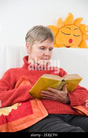Upper body view of an elderly woman reading a book on a white sofa. Behind her a sun-shaped sofa cushion on a white wall. Stock Photo