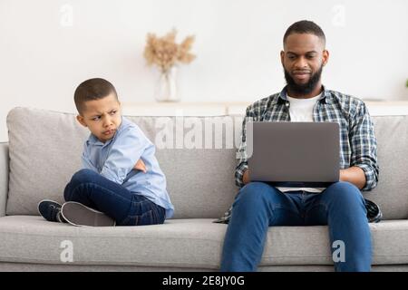 Indifferent African Father Using Laptop Ignoring Offended Son At Home Stock Photo