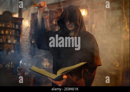 Male exorcist in black hood holds book of spells Stock Photo