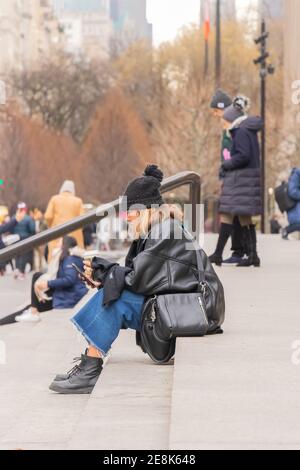 young woman in woolly hat looks at cell phone while sitting on stone steps in new york on steps of New York The Metropolitan Museum of Art Stock Photo