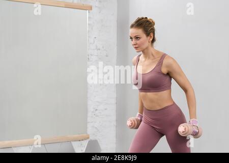 Girl in tight pink outfit working out with dumbbells. Beautiful fitness girl training legs and lifting weights. Girl doing sports in a loft interior Stock Photo