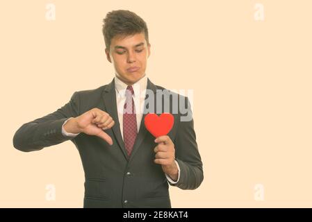 Young sad businessman giving thumb down while holding and looking at red heart ready for Valentine's day Stock Photo