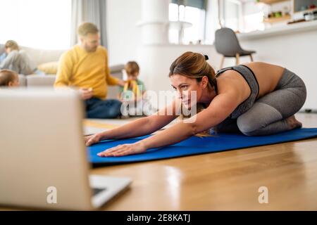 Young woman, mother exercising at home in living room, father playing with kids in background. Stock Photo