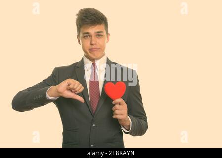 Young sad businessman giving thumb down while holding red heart ready for Valentine's day Stock Photo