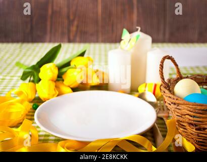 Easter table setting with white plate, basket of colorful eggs, candles and yellow tulips Stock Photo
