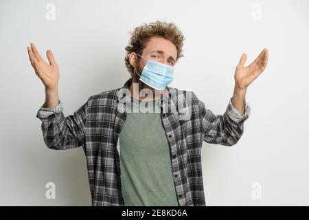 Young man in protective face mask with curly hair showing I don't know gesture with both hands isolated on white background. Portrait of confused sick Stock Photo