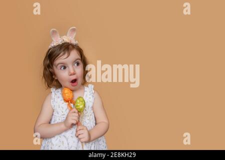 Positive portrait of little girl with rabbit ears on head, looks in surprise, holding an orange green egg in her hands. Isolated light orange backgrou Stock Photo