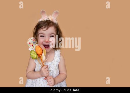 Emotional, positive portrait of little girl with bunny ears on head holding an Easter bouquet of eggs, chickens. Isolated light orange background plac Stock Photo