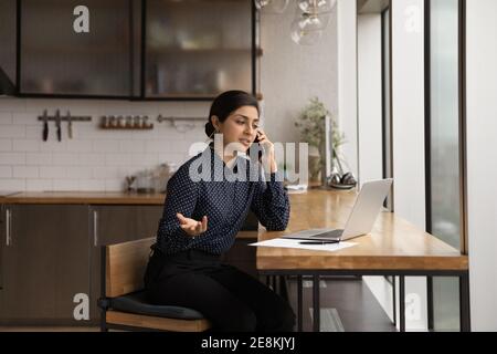 Indian female talk by cell reading documents on laptop screen Stock Photo