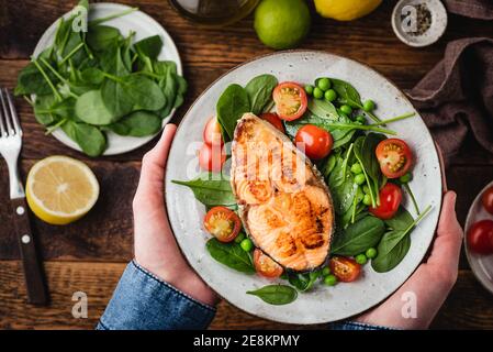 Healthy dinner meal with roasted salmon steak, baby spinach and cherry tomatoes salad. Male hands holding plate with healthy food Stock Photo