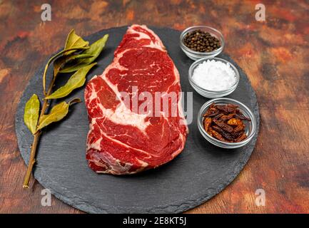 Raw beef ribeye on a round slate plate with glass containers containing peppercorns, coarse salt and cayenne chillies on an old wooden table in brown Stock Photo
