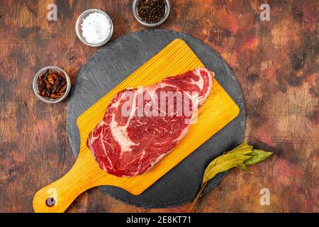Raw beef ribeye on a round slate plate with glass containers containing peppercorns, coarse salt and cayenne chillies on an old wooden table in brown Stock Photo