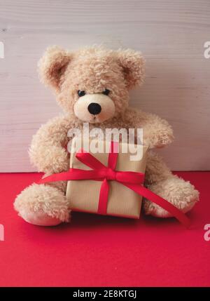 Valentines day, Christmas present. Teddy bear holding a gift box sitting on red floor, wooden wall background, copy space, Holiday birthday greeting c Stock Photo