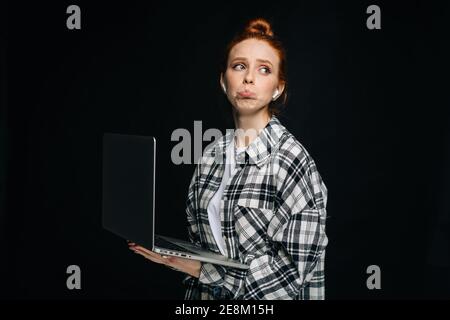 Sad upset young woman wearing wireless earphones holding laptop computer and looking at camera. Stock Photo