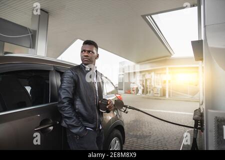 Gas petrol station concept. Relaxed dreamy young handsome African businessman in black outfit, enjoying his take away coffee, while refueling his Stock Photo