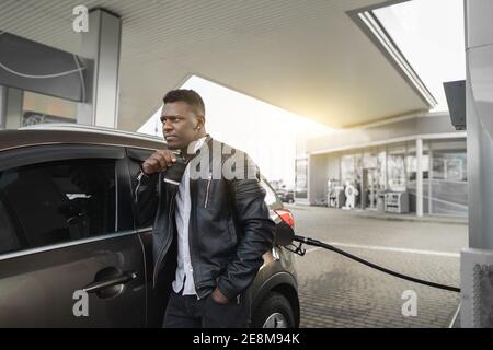 Gas petrol station concept. Relaxed dreamy young handsome African businessman in black outfit, enjoying his take away coffee, while refueling his Stock Photo