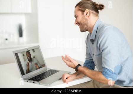 Young handsome hipster guy sitting at the desk at the home office and talking to virtual African American friend on webcam, telling a joke, or Stock Photo