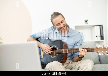 Attractive joyful bearded hipster man smiling and playing the guitar, sitting in the living room on a comfy couch, learning a song from online music Stock Photo