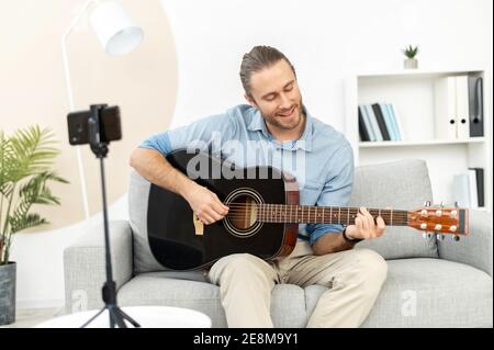 Handsome attractive bearded hipster guy making video blog about musical instruments, sitting on the couch, playing the guitar, and streaming tutorial Stock Photo