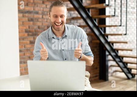 Positive young male entrepreneur approves the project online on the laptop, looking at the screen and showing thumbs-up, moved into a new modern house Stock Photo