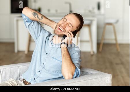 Stylish young bearded hipster guy is chilling at home, sitting on the comfy sofa in the modern living room, stretching his neck and holding a mobile Stock Photo