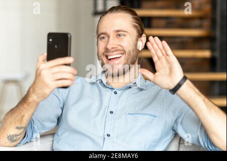 Smiling bearded hipster man sitting on the couch in the living room, video-calling, talking to friend, family, colleague, waving at the mobile phone Stock Photo
