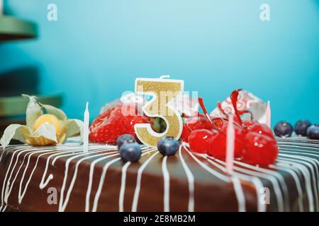 Birthday-anniversary cake with gold candle showing number 3.Chokolate cake with fruits and a candle with the number three on blue background Stock Photo