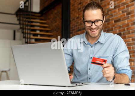 Young bearded businessman wearing glasses using laptop and credit card for online payment at home, disabled guy in wheelchair holding the debit card Stock Photo