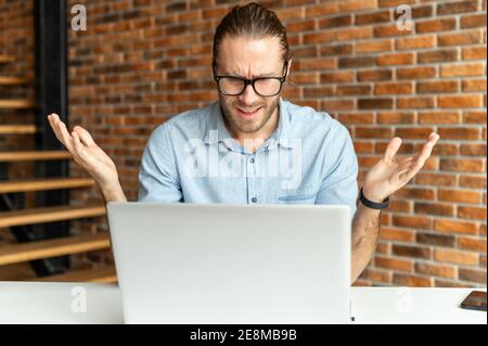 Young hipster guy in glasses sitting at the desk in front of a laptop and raising his hands up, feeling frustrated and scared from occurred issues Stock Photo