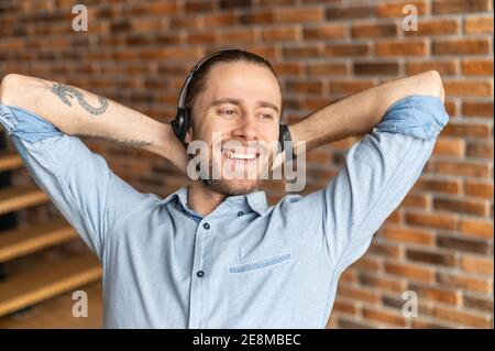 Millennial young male freelancer in headset placed his hands behind head, hipster customer service representative posing against the brick wall Stock Photo