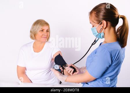 A young female doctor in a medical uniform and disposable mask measures the blood pressure of an caucasian senior woman on a white background. Stock Photo